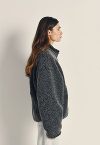 Szara, puszysta kurtka polarowa z wysokim kołnierzem i szerokimi rękawami, z teksturowanym wykończeniem i zapinanym na zamek dekoltem, noszona na jasnym dżinsie.