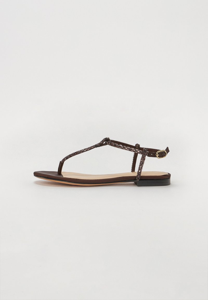 Sandali piatti marroni con design a T-strap, caratterizzati da dettagli intrecciati e una piccola fibbia alla caviglia. La suola è di un marrone scuro a contrasto.