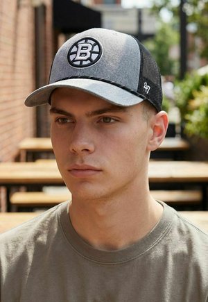 Junger Mann trägt eine graue und schwarze Baseballkappe mit einem "B"-Logo, sitzt tagsüber an hölzernen Tischen im Freien.