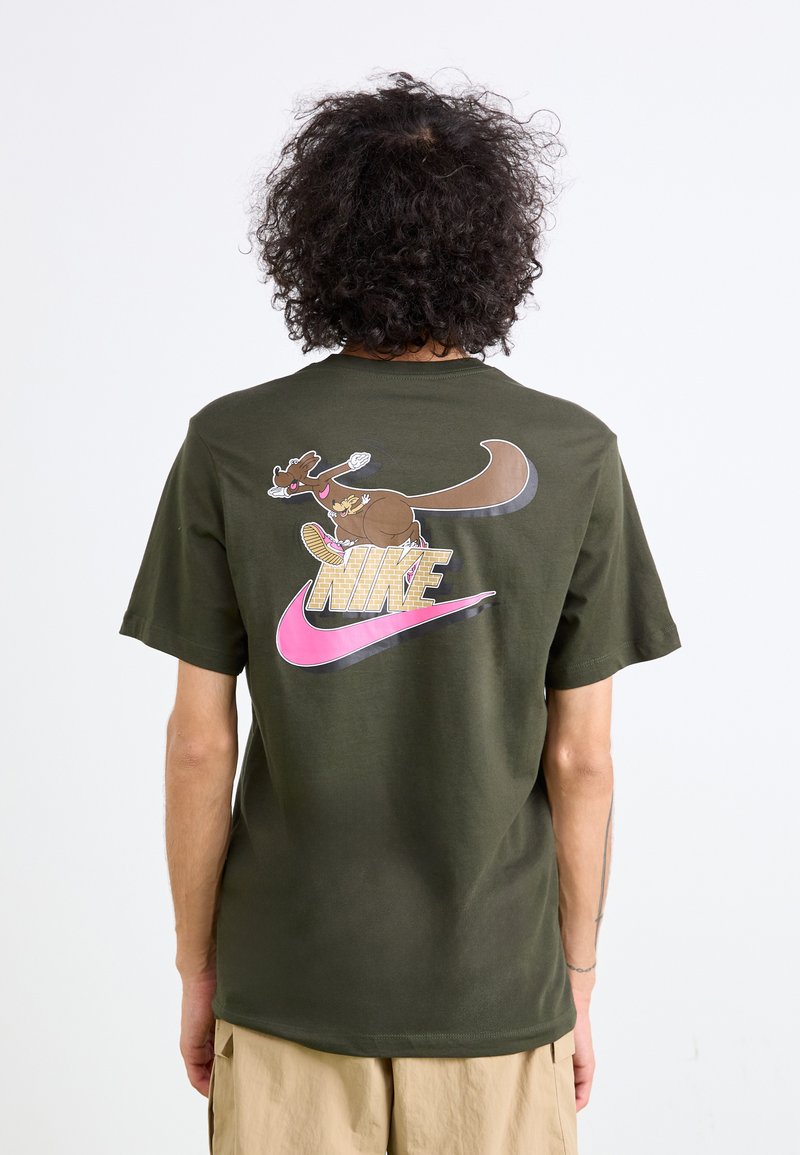 Olivengrøn bomulds t-shirt med et grafisk design af et tegneserieagtigt dyr iført en sneakers og et stort pink Nike-logo på ryggen.