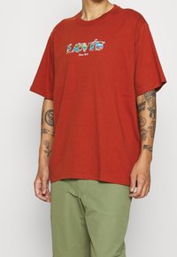 T-shirt en coton rouge, coupe ample, col rond, avec un graphisme coloré "Levi's Since 1873" sur le devant. Porté avec un pantalon vert.