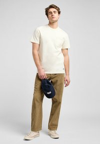 Light cream cotton t-shirt with pocket, paired with beige corduroy pants. Model holds a navy cap. Shoes are white canvas with red accents.