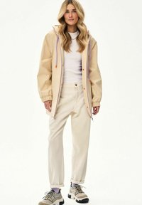 Kobieta o długich, falowanych blond włosach, ubrana w beżową kurtkę z kapturem, białą koszulkę, kremowe spodnie i beżowe masywne trampki, stojąca na białym tle.