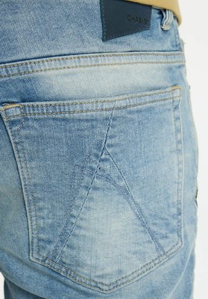 Svetlomodré denimové rifle s výrazným dizajnom zadného vrecka a žltým prešitím. Majú tmavú koženú značkovú náplasť na páse.