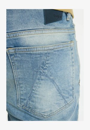 Hele sinised teksapüksid, millel on iseloomulik tagatasku disain ja kollane õmblus. Vöökohal on tume nahast kaubamärgi märk.