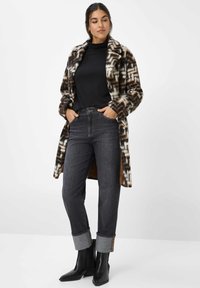 Frau, die einen schwarzen Rollkragenpullover, einen gemusterten braun-weißen Mantel, dunkle hochgekrempelte Jeans und schwarze Stiefeletten trägt, steht mit den Händen in den Taschen.