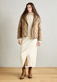 Veste matelassée brune rembourrée, pull crème en tricot torsadé et jupe crème avec une fente latérale ; bottes en cuir à lacets marron. Palette de couleurs neutres.