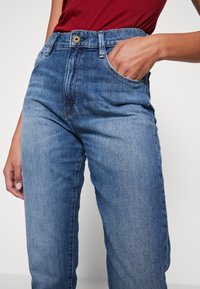 Light blue denim jeans with a high waist, straight leg design, visible front pockets, and a gold button at the waistband.