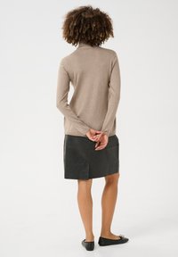 Beige garbó pulóver, slim-fit, hosszú ujjú; fekete bőrszoknyával párosítva. A modellen keresztbe tett karokkal áll, fekete lapos cipőt visel.