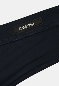 Bañador corto de color azul marino con una tela texturizada y una etiqueta rectangular negra con la marca "Calvin Klein" en blanco en la cintura.