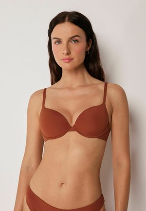 Frau mit langem dunklem Haar, die einen rostfarbenen BH und passende Unterwäsche trägt und vor einem schlichten hellen Hintergrund steht.