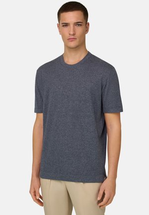 Jeune homme aux cheveux courts portant un t-shirt à manches courtes gris foncé uni et un pantalon beige, debout devant un fond blanc.