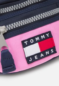 Borsa Tommy Jeans con corpo in tessuto rosa, accenti blu navy e cerniere in metallo, caratterizzata da un'etichetta con il logo nei colori blu navy, bianco e rosso.