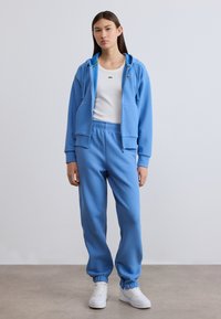 Φωτεινό μπλε φούτερ με φερμουάρ και ασορτί παντελόνι φόρμας με ελαστικές μανσέτες, φορεμένα πάνω από λευκή αμάνικη μπλούζα. Διαθέτει λογότυπο στο στήθος.
