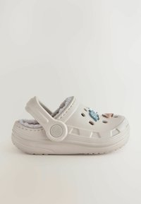 Sabots blancs avec un intérieur en peluche, présentant des charms décoratifs colorés et des trous de ventilation, conçus pour le confort et la facilité à porter.