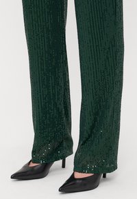 Roheline sädelusega püksid, millel on lõtv lõige ja tekstureeritud pind. Sobitatud teravate mustade kõrge kontsaga kingadega, mis rõhutavad riietust.