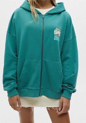 Femme portant un sweat à capuche zippé vert sarcelle avec poches avant et un dessin en texte blanc sur la poitrine, par-dessus une jupe blanche effilochée.