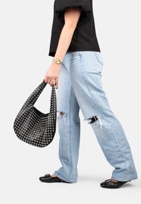 Bolso hobo negro con tachuelas y un diseño holgado. Lleva unos vaqueros claros desgastados con grandes desgastes. Zapatos planos negros con un detalle de hebilla.