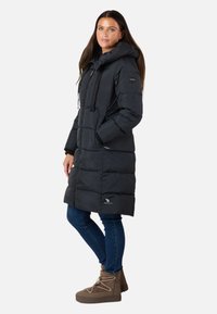 Cappotto invernale imbottito nero con cappuccio, dotato di zip, tasche e polsini, abbinato a jeans blu e stivaletti alla caviglia marroni.