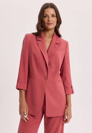 Kobieta z falującymi brązowymi włosami ubrana w dopasowaną różowawą marynarkę z podwiniętymi rękawami i pasujące spodnie na jednolitym jasnym tle.