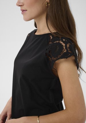 Femme portant un haut noir avec des manches courtes en dentelle, des boucles d'oreilles créoles dorées, un collier fin et un bracelet en or au poignet gauche.