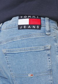 Ljusblå denimjeans med en texturerad yta. Har en svart etikett med "TOMMY JEANS" och röda, vita och blå detaljer.