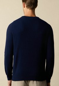 Granatowy sweter, długie rękawy, okrągły dekolt, ściągacze przy rękawach i u dołu, miękka tekstura, minimalistyczny design, noszony na jasnych spodniach.