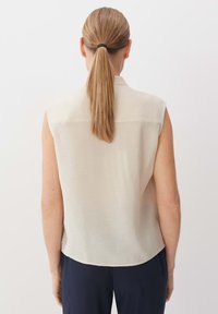 Blusa senza maniche di un beige chiaro con cucitura posteriore e orlo arrotondato. Realizzata in un tessuto liscio e leggero. Nessun motivo o accento visibile.