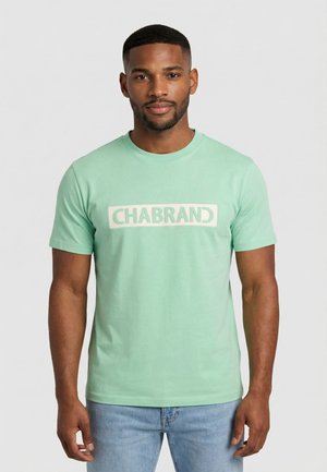 Uomo con barba corta che indossa una maglietta verde menta con il logo "CHABRAND" e jeans azzurri, in piedi davanti a uno sfondo grigio uniforme.