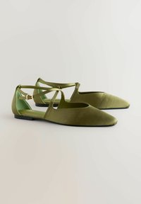 Zapatos planos de color verde oliva con punteras puntiagudas, correas entrelazadas y un acabado de material suave. Incluyen un detalle de hebilla cerca del talón.