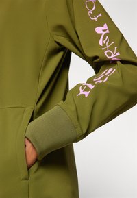 Rukav sweatshirt boje masline s ružičastim grafičkim tekstom i rebrastim manžetama, ruka djelomično u bočnoj džepu na jednostavnoj pozadini.