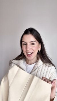 Leende kvinna med långt mörkt hår håller en vikt krämfärgad tröja, klädd i en vit stickad jacka med dragkedja och pärlörhängen.