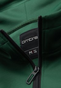 Veste vert foncé avec fermeture éclair noire partiellement ouverte montrant la marque "ombre" et l'étiquette taille M (EUR 48) à l'intérieur du col.