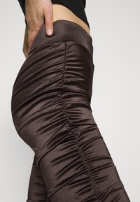 Leggings marron froncés en tissu brillant, dotés d'une taille haute et de coutures verticales texturées pour un design ajusté.