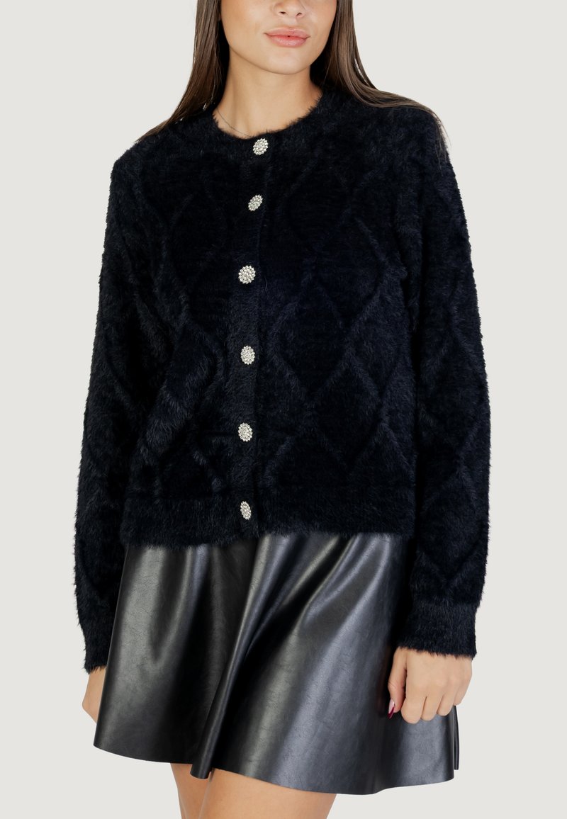 Cardigan en laine bleu marine, moelleux, avec un motif en losanges sur le devant, col rond et six fermetures à boutons argentés, associé à une jupe en cuir noire.