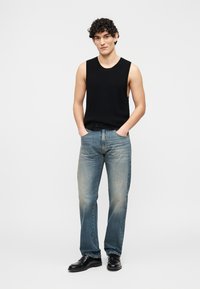 Jeune homme aux cheveux bouclés portant un haut noir sans manches, un jean bleu délavé et des chaussures habillées noires, les mains dans les poches.