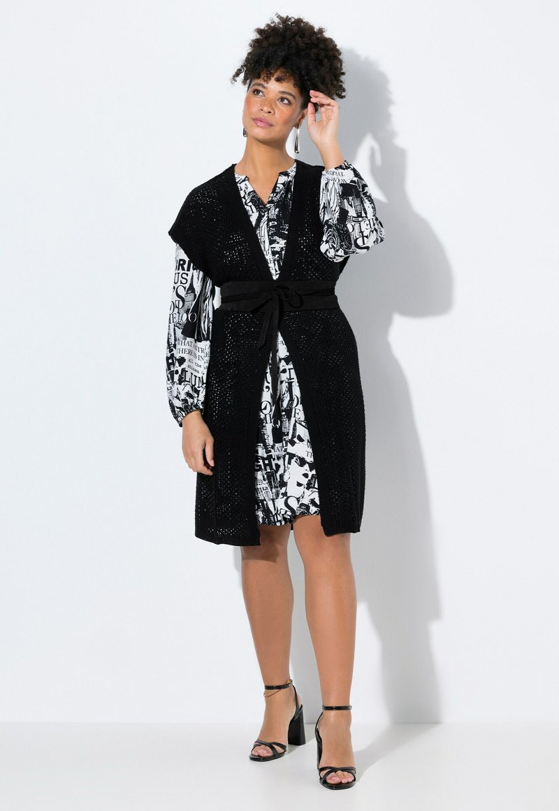 Gilet long en tricot noir avec devant ouvert, ceinturé à la taille. En dessous, une robe à manches longues noir et blanc à motifs. Sandales à talons hauts.