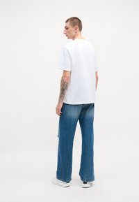 Camiseta de algodón blanca de mangas cortas combinada con vaqueros de pierna ancha azules y zapatillas deportivas blancas. Tatuaje visible en el antebrazo izquierdo.