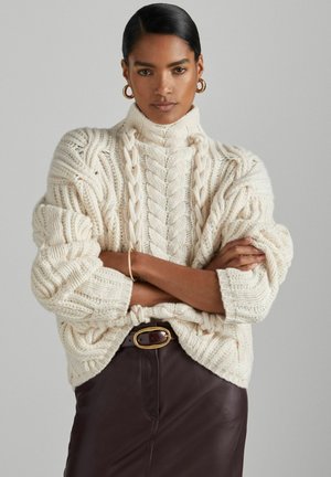 Chunky cream cable-knit sweater with high collar, featuring thick textured stitching. Worn over a dark leather skirt with a belt.