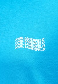 Φωτεινό μπλε ύφασμα με την επιγραφή "KARL LAGERFELD" να επαναλαμβάνεται τέσσερις φορές σε λευκό κυματιστό κεφαλαίο κείμενο στο κέντρο της επιφάνειας.