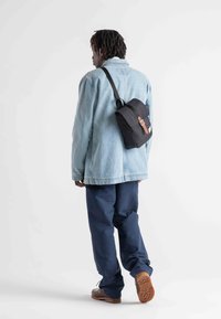 Black shoulder bag with a flap, featuring a leather strap and a logo tag, worn over a light denim jacket and navy pants. Brown footwear visible.
