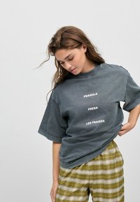 Camiseta gris de gran tamaño con texto blanco que dice "FRAGOLA," "FRESA," y "LES FRAISES." Combinada con pantalones de cuadros en verde oliva.