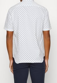 Camicia bianca a maniche corte con un piccolo motivo a pois blu navy, colletto button-down e tessuto traspirante. Presenta un orlo dritto e spacchi laterali.