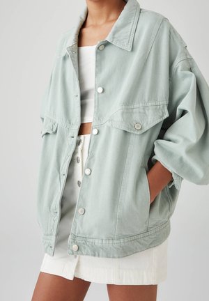 Personne portant une veste en jean oversize vert clair sur un crop top blanc et une jupe blanche boutonnée, la main dans la poche de la veste.