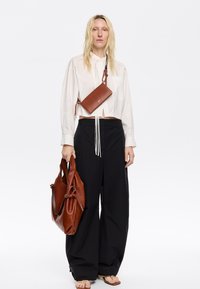 Borsa a tracolla in pelle marrone con dettagli intricati, abbinata a una camicia bianca con colletto e pantaloni black a gamba larga. Sandali neutri completano il look.