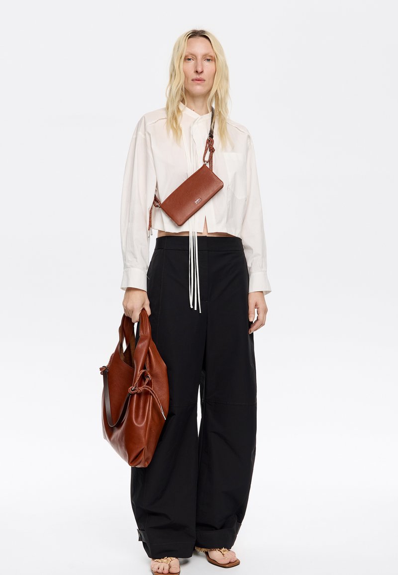 Borsa a tracolla in pelle marrone con dettagli intricati, abbinata a una camicia bianca con colletto e pantaloni black a gamba larga. Sandali neutri completano il look.