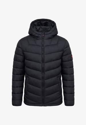 Teenager-Junge steht mit den Händen in den Taschen, trägt eine schwarze gesteppte Jacke, schwarze Hosen mit Reißverschlüssen und schwarze Sneakers vor grauem Hintergrund.