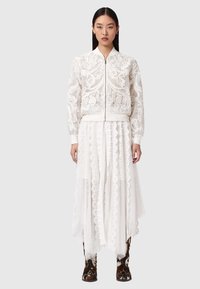 Ung kvinde iført en hvid, gennemsigtig broderet bomberjakke, lang blonde nederdel og brune mønstrede støvler, stående mod en ensartet baggrund.