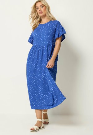 Blondynka w niebieskiej sukience midi w białe grochy z falbankowymi krótkimi rękawami, trzyma sukienkę, stojąc w białych sandałach.