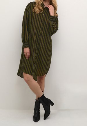 Kobieta stojąca, ubrana w zielono-czarną, pionowo prążkowaną sukienkę na guziki, zestawioną z czarnymi botkami na obcasie.